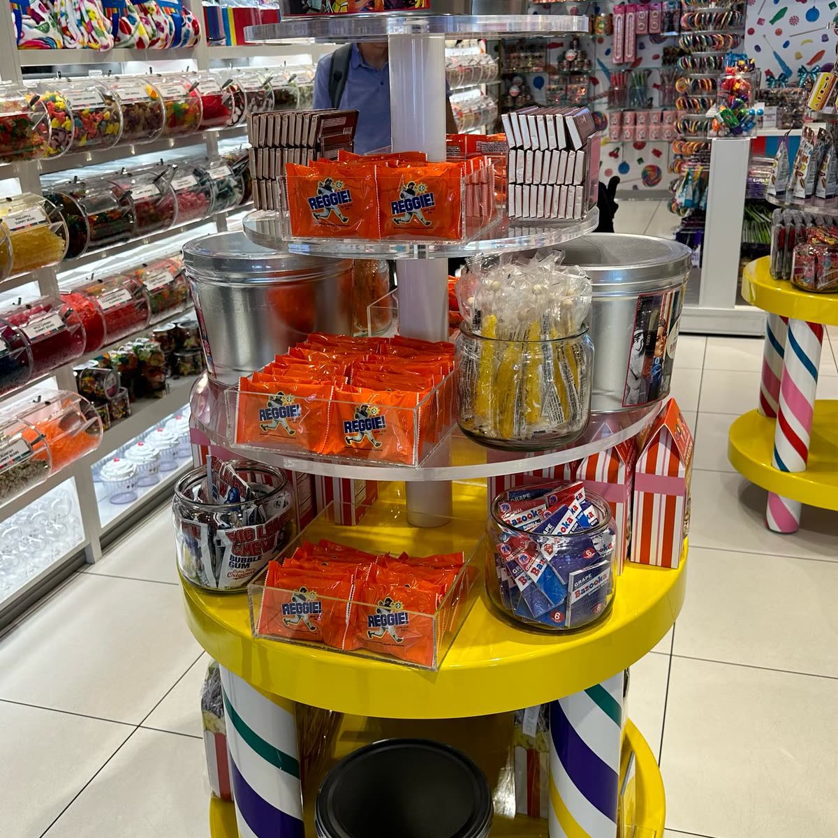 A tiered candy store display filled with trays of orange-wrapped REGGIE! Bars