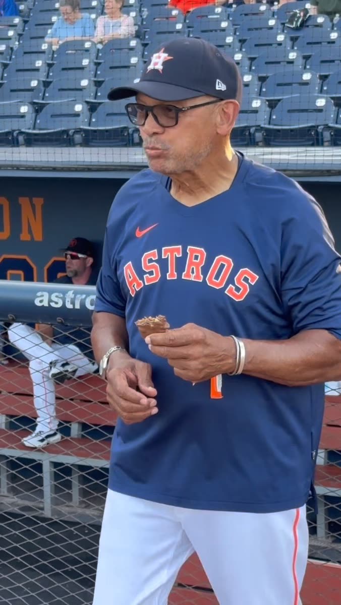Reggie Jackson in a baseball jersey and cap eating a REGGIE! Bar beside the dugout railing